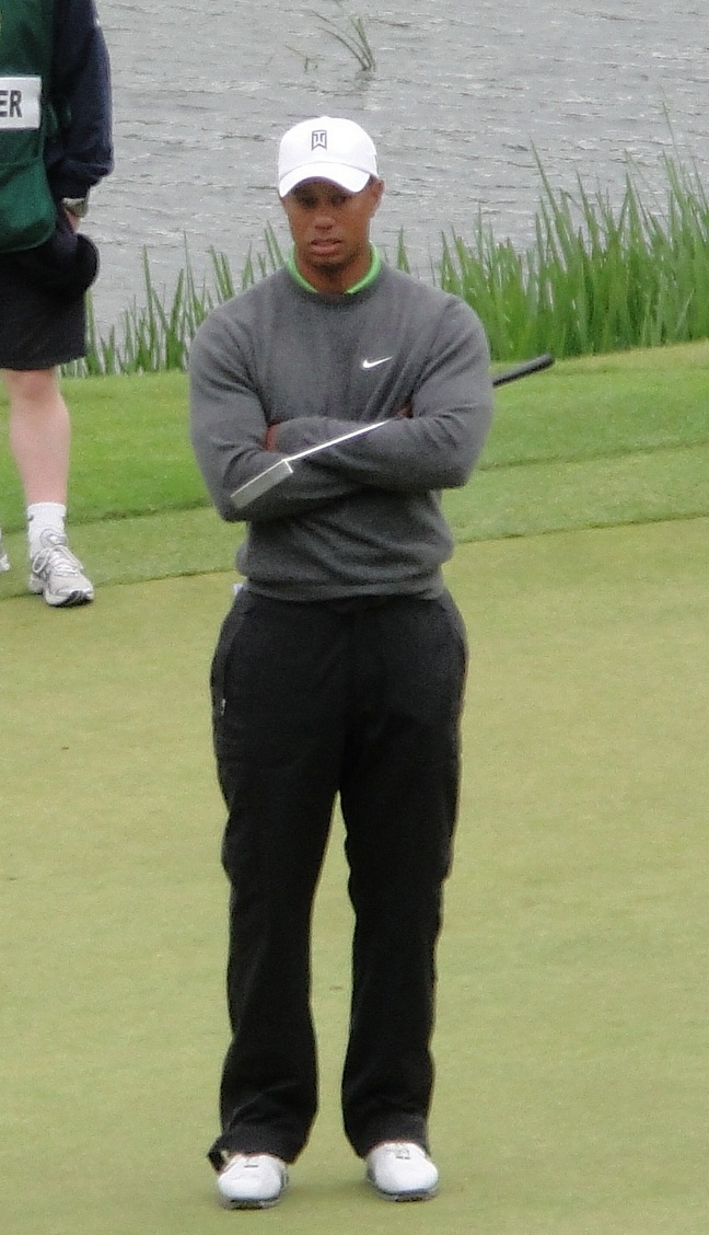 Tiger Woods, black pants, gray nike sweater, white golf shoes, white baseball hat